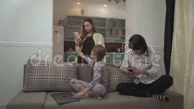 两个姐姐在手机上发短信，小女孩坐在房间里的沙发上用笔记本电脑打字。 小可爱